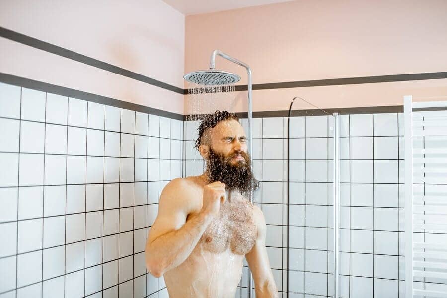 Bearded man feeling shocked while taking a shower with cold water in the bathroom at home