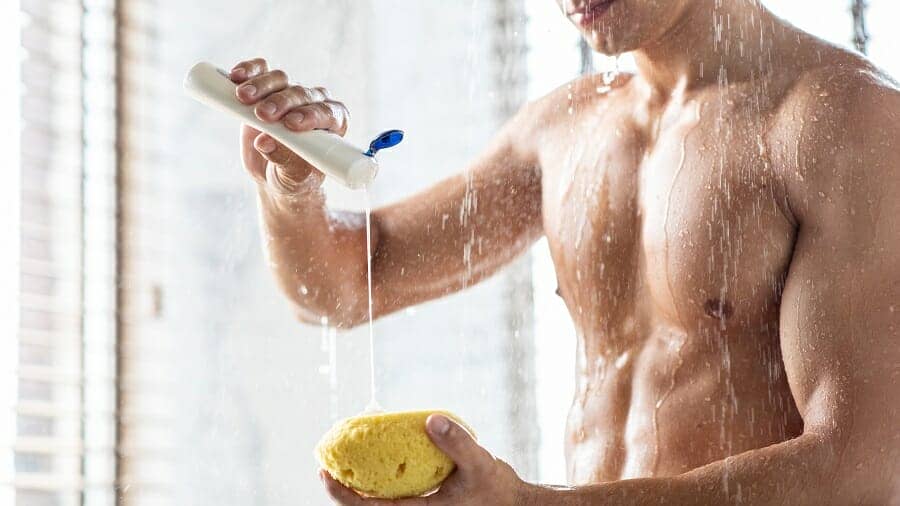 Man die onder de douche staat en shampoo op gele spons giet